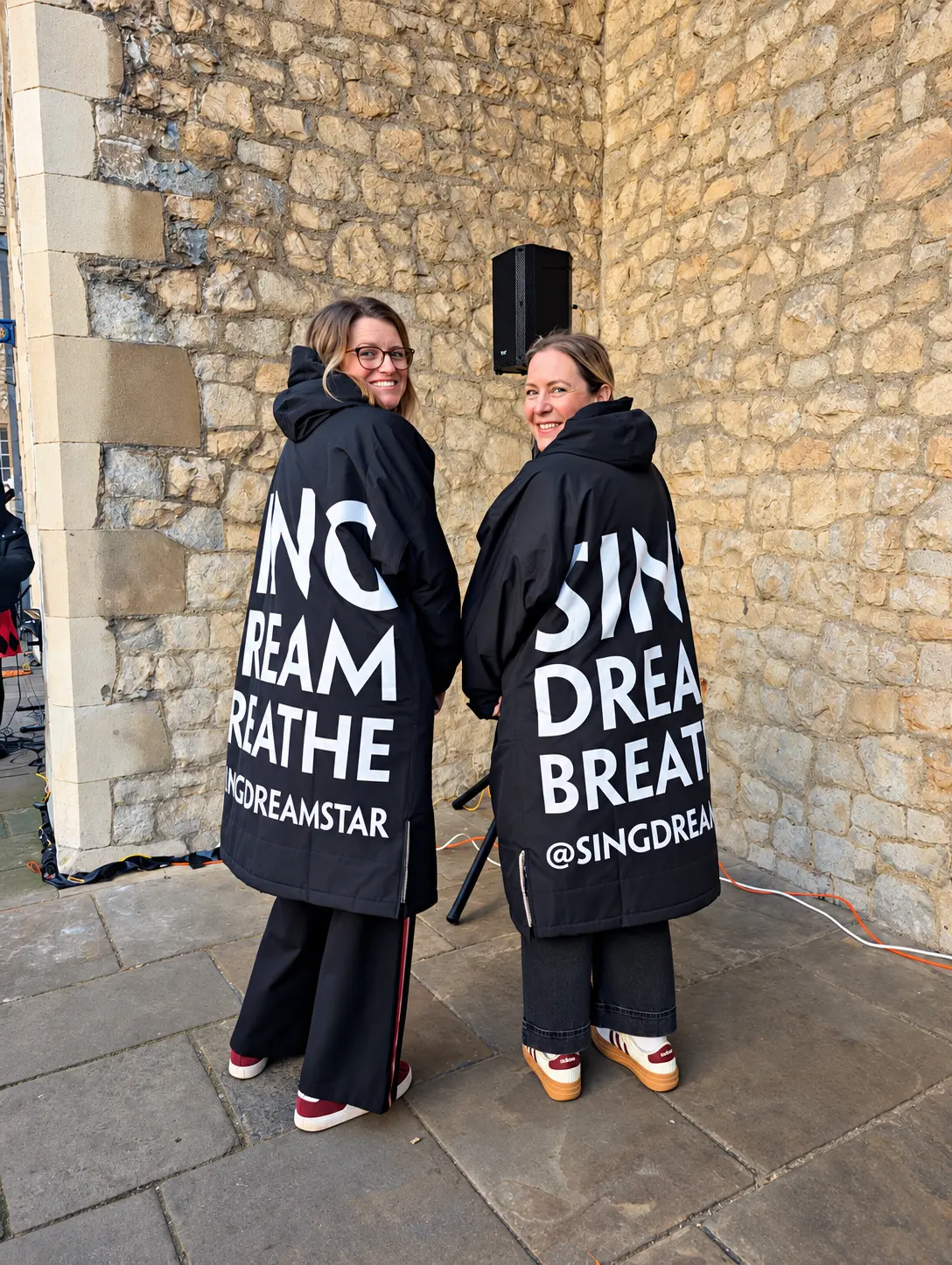 Two choir members wearing Sing Dream Breathe coats beside a stone wall