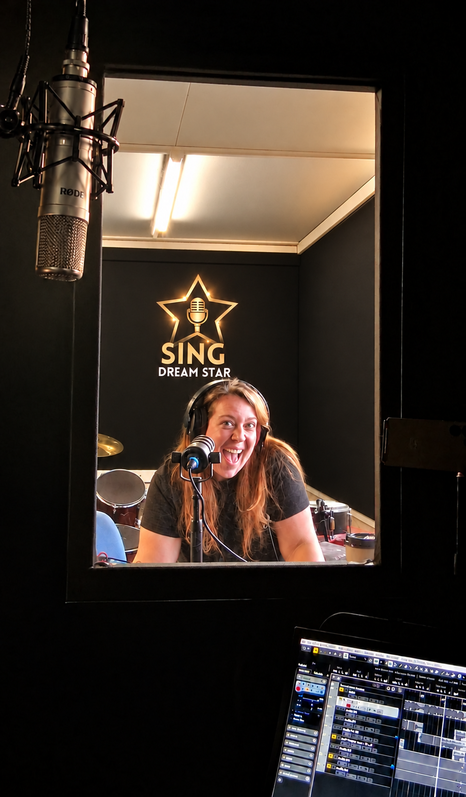 A smiling singer wearing headphones at a microphone through the recording studio window